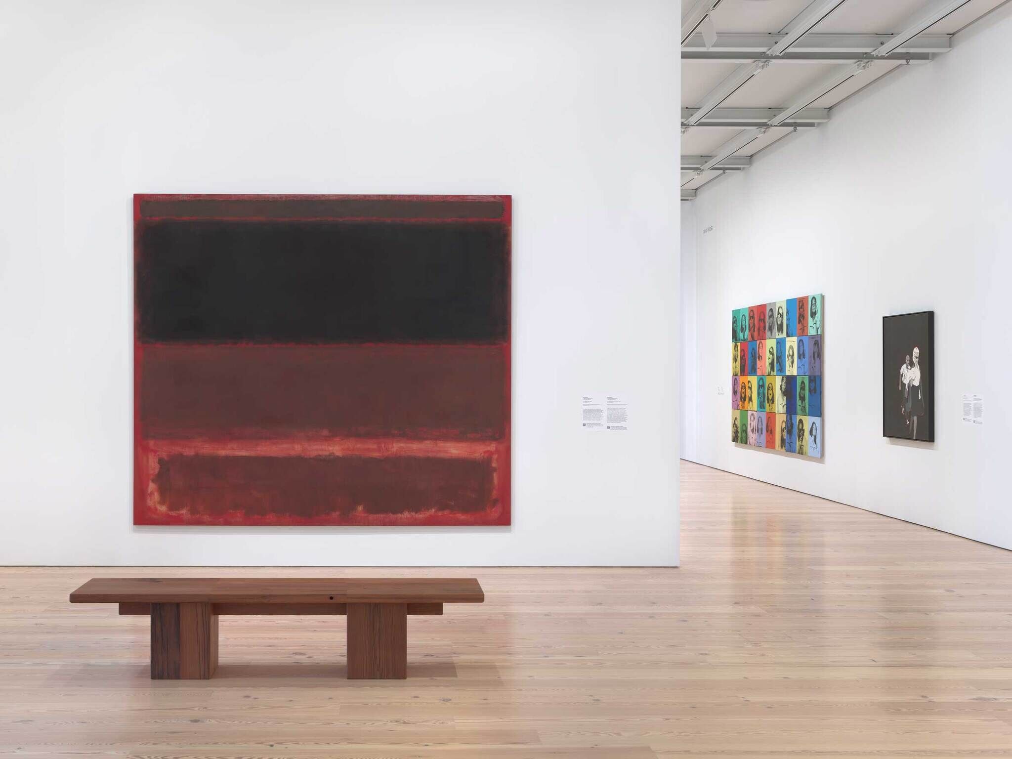 Large abstract painting with dark and red blocks on a white wall, wooden bench in front, other colorful art nearby.