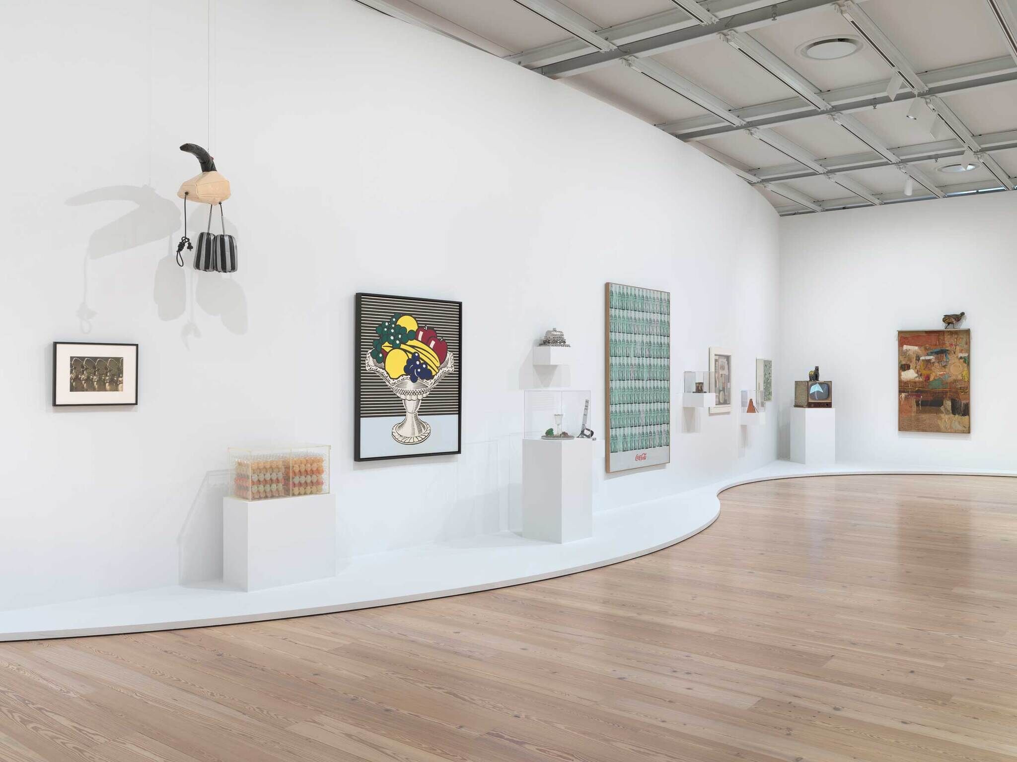 Modern art gallery with various paintings, sculptures, and objects displayed along a curved white wall and wooden floor.