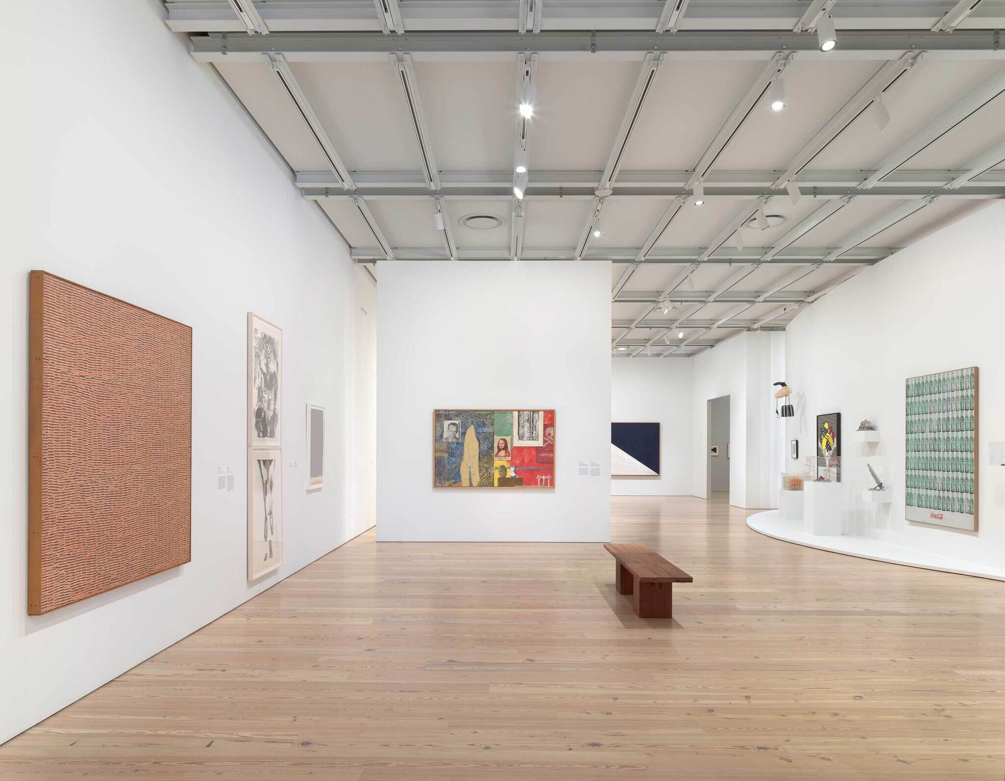 Art gallery with modern paintings and sculptures on white walls, wooden floor, and a wooden bench in the center.