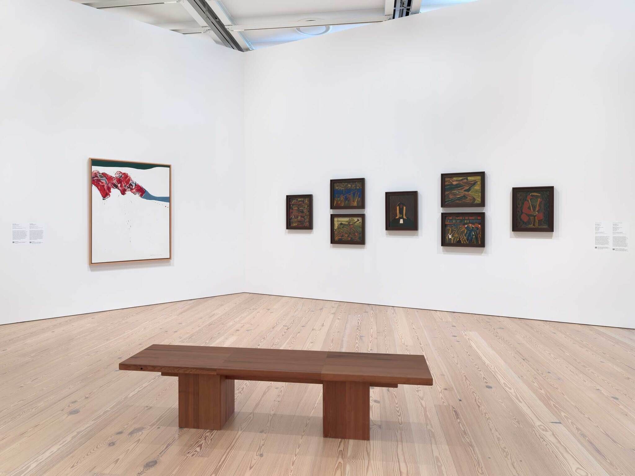 A wooden bench sits in front of several framed paintings displayed on white gallery walls.