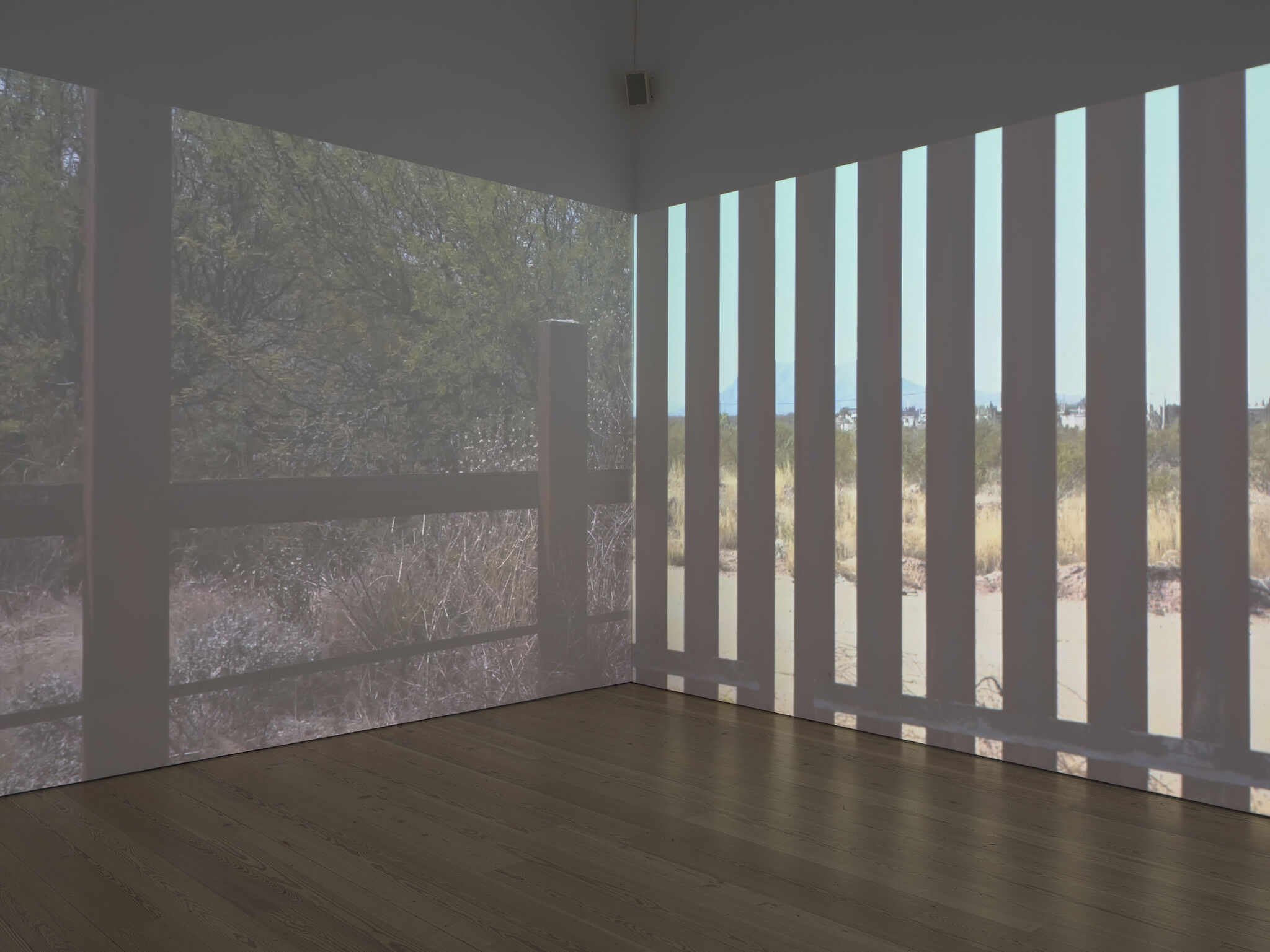 A video installation projected on two walls that meet in a corner. The images are of landscapes seen through a fence.