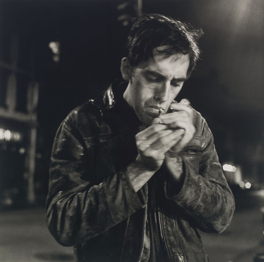 A man in a leather jacket lights a cigarette on a dimly lit street at night.